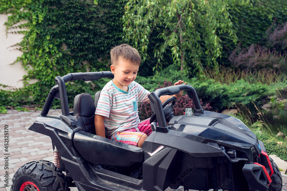 Smiling excited boy of 5 years old sits in black toy electrocar with green fence on the background. Driving child