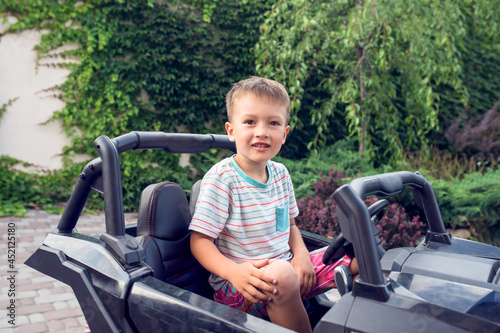 Excited little boy sits in a black electrocar in front of green fence and a tree. Holidays and free time for preschoolers