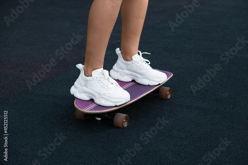 close-up of the female legs of a Caucasian girl, shod in white women's sneakers, standing on a purple skateboard in the park against the background of a treadmill