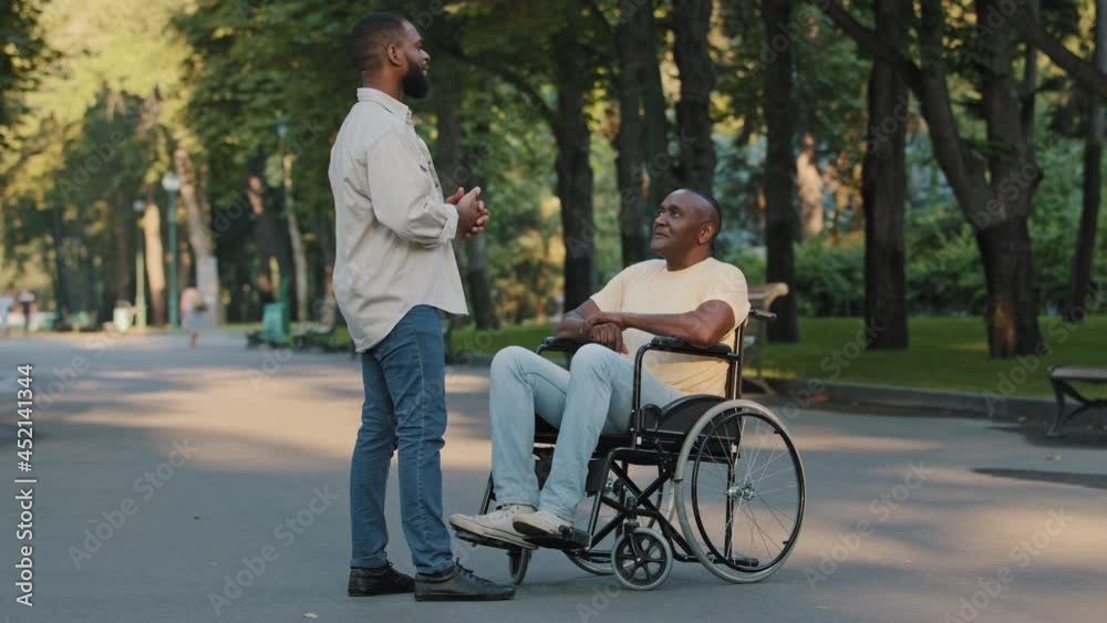 Two African men relaxing in hospital park, having fun outdoors, chatting talking about championship. Black disabled man in wheelchair gesturing vigorously while discussing basketball game with friend Two African men relaxing in hospital park, having fun outdoors, chatting talking about championship. Black disabled man in wheelchair gesturing vigorously while discussing basketball game with friend