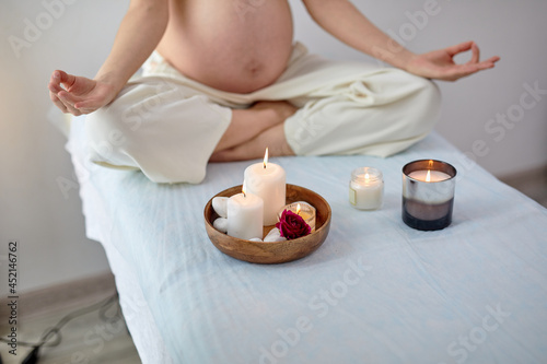 Cropped pregnant woman sitting in yoga position in spa room, focus on candles, relaxing and meditating while wearing white outfit, showing naked belly. Health lifestyle pregnancy beauty, interior.