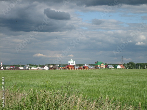 View of a green field overgrown with grass. Behind the field, you can see a village with a towering bell tower of an Orthodox church.