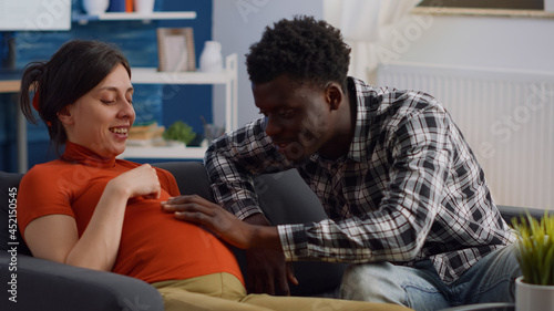 Wallpaper Mural Pregnant interracial couple sitting on living room couch. African american husband touching baby bump and talking to child while caucasian mother relaxing on sofa and laughing Torontodigital.ca