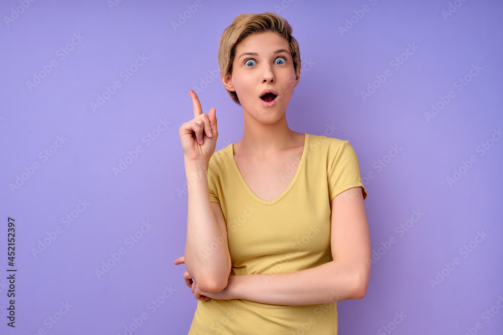 Fototapeta premium Portrait Of Cheerful Female In Yellow Casual T-shirt Having Great Brilliant Idea, Standing With Wide Opened Mouth, Raising Finger Up, Thinking Creating New Plan, Isolated On Purple Background