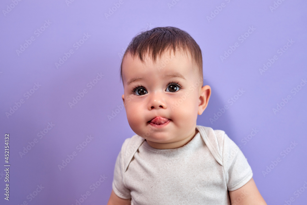 Cute Caucasian Baby Showing Tongue At Camera, Looking Sweet, Isolated On Purple Studio Background, Portrait of Little Kid In Casual T-shirt Looking At Side, With Amazed Facial Expression