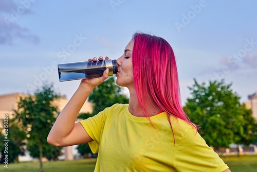 cute young woman with pink hair drinks water from a reusable bottle after training outdoors