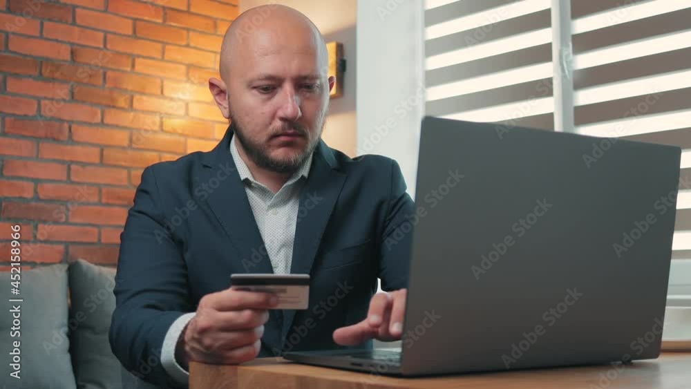 Young man in the suit holding a credit card and making online purchases through a laptop, paying at online store, e-commerce concept