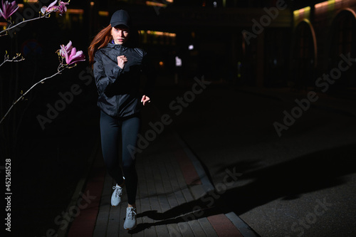 Attractive Young Fitness Woman in Black Athletic Wear Outfit Jogging On Street Alone, Night. Caucasian Slim Slender Female Is Doing Workout in Night Urban Environment, Running Forward. Copy Space © alfa27