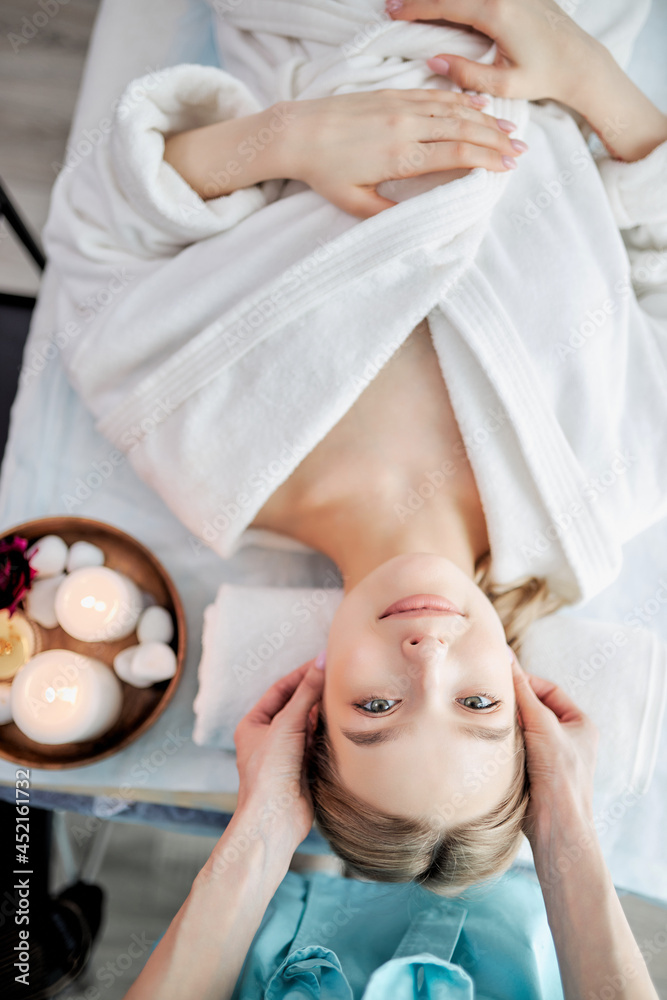 Top view of positive woman relaxing while careful specialist massaging her face, adorable blonde lady in white bathrobe lying on couch enjoying spa procedure, cropped masseur hands massaging face Top view of positive woman relaxing while careful specialist massaging her face, adorable blonde lady in white bathrobe lying on couch enjoying spa procedure, cropped masseur hands massaging face