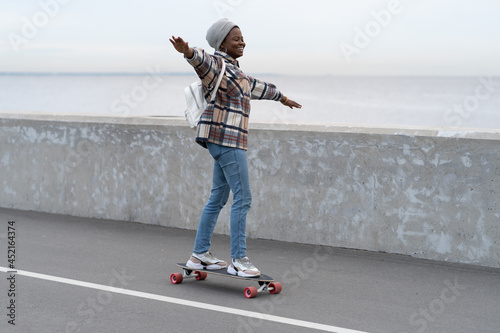 Freedom and urban lifestyle concept: young casual girl skateboarding on longboard at city road near sea coast. Happy millennial active african american woman enjoy extreme leisure activity outdoors. photo