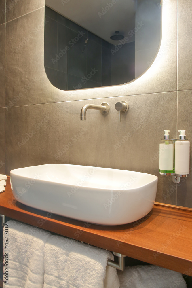 Fototapeta premium Modern simple bathroom with white bathroom sink standing on wooden shelf and backlit mirror