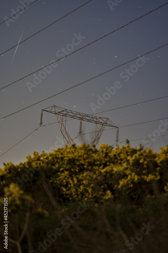 Beautiful night view of power lines, electric transmission pylon, falling Lyrid meteor and spring yellow flowering gorse (Ulex) in Ticknock, County Dublin, Ireland. Soft and selective focus