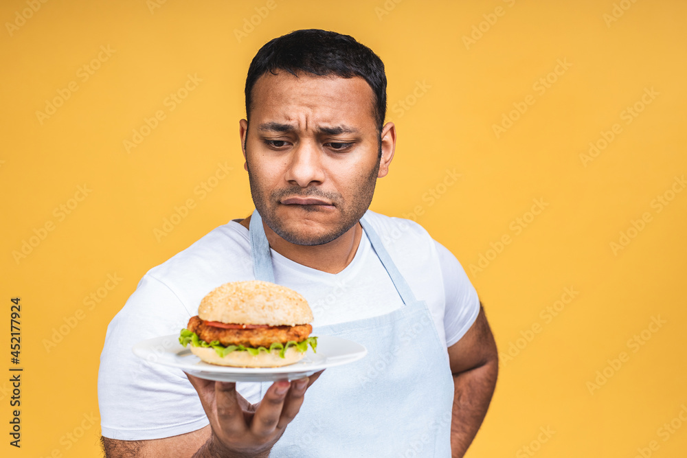 Young african american indian black man eating hamburger isolated over yellow background. Cook preparing burger. Young african american indian black man eating hamburger isolated over yellow background. Cook preparing burger.