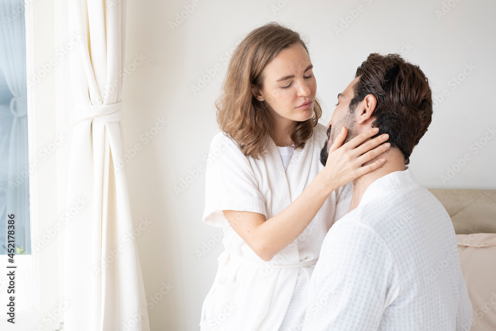 Romantic couple cuddling while sitting in bed in the morning. Happy couple sitting on bed and kissing each other at home. Happy young couple on bed, female come and embrace male.