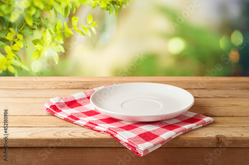 Empty white plate on wooden table with red checked tablecloth over  blurred nature background.  Kitchen mock up for design and product display.