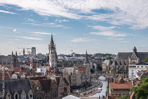 Gent, Flanders, Belgium - July 30, 2021: Aerial view on medieval Korenlei along Leie River with Postal clock tower and Saint Michael church. Cityscape with cranes under blue cloudscape. © Klodien