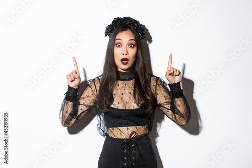 Impressed asian girl in black lace dress and wreat turn your attention on halloween promo, making announcement, pointing fingers up, showing banner, standing over white background photo