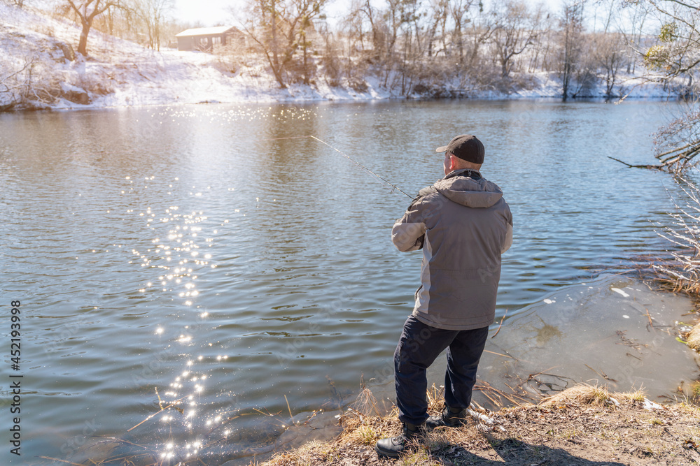 Fototapeta premium A fisherman with a fishing rod catches fish on the banks of a snow-covered river in early spring
