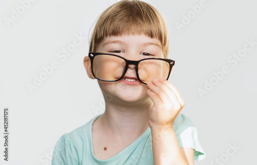 A cheerful little girl with glasses on her nose on a light background. A contented child with glasses. A joyful little girl with glasses smiles, feeling happy. Children's games, pampering. Soft focus