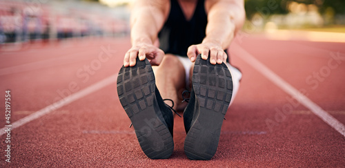 Young muscular athletic runner man stretching and touching his feet on a running court in sitting position before starting of running - Jogging and flexibility concept - Focus on the runner's shoes