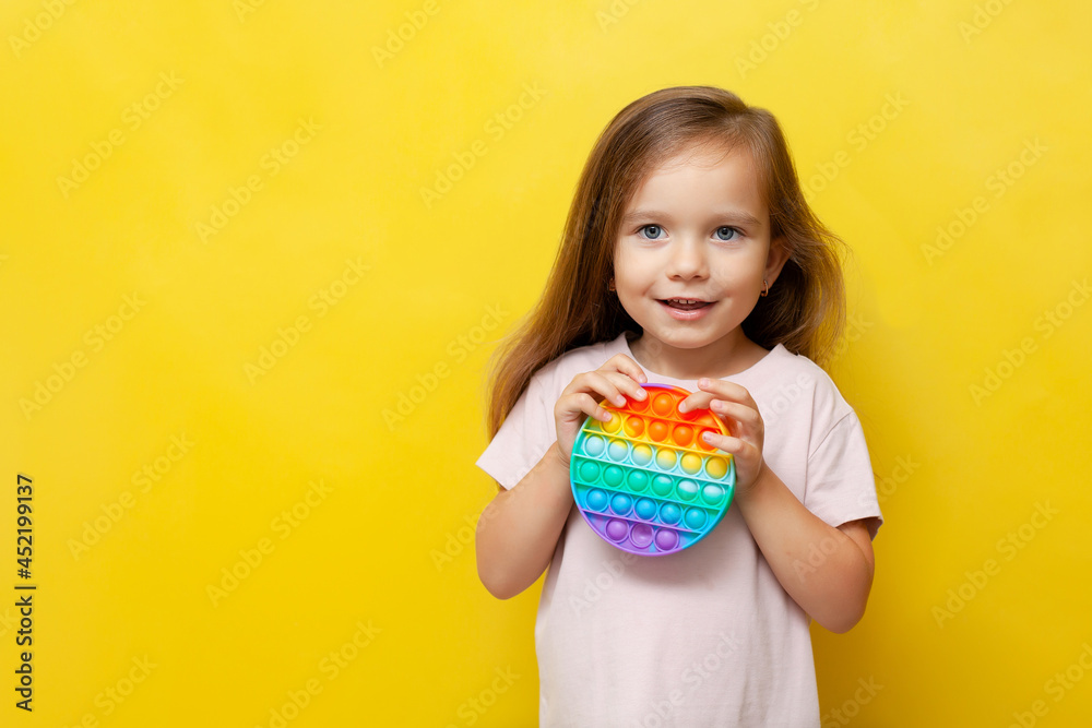 A beautiful cute girl holds a colored pop it toy in her hands and smiles. Yellow background. Anti-stress, emotions, good mood. A place for text. Trend A beautiful cute girl holds a colored pop it toy in her hands and smiles. Yellow background. Anti-stress, emotions, good mood. A place for text. Trend