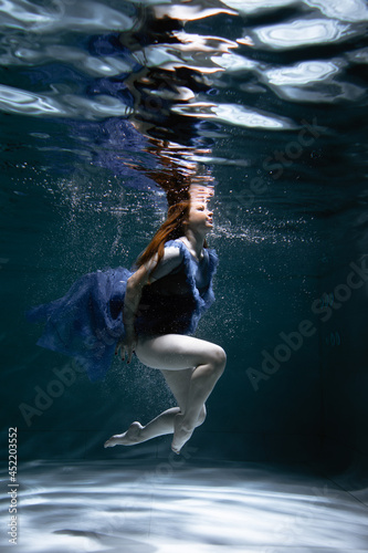 A beautiful girl in a blue dress swims underwater in the pool like a little mermaid from a fairy tale