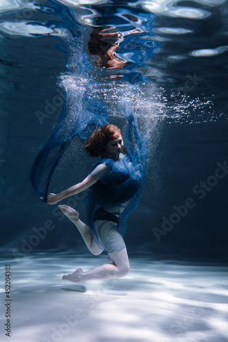 A beautiful girl in a blue dress swims underwater in the pool like a little mermaid from a fairy tale