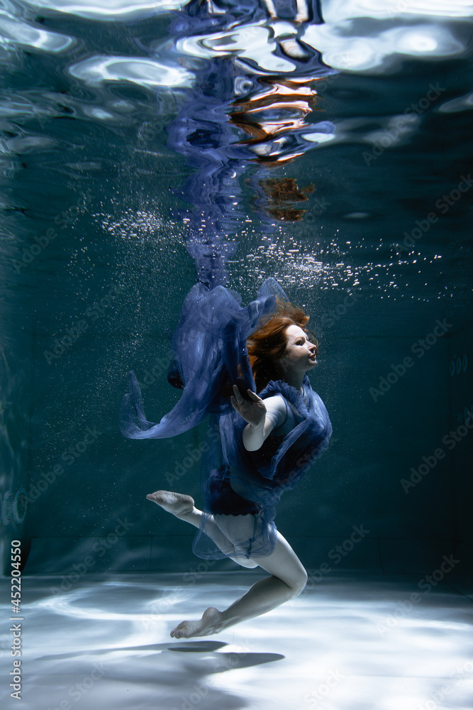 Fototapeta premium A beautiful girl in a blue dress swims underwater in the pool like a little mermaid from a fairy tale