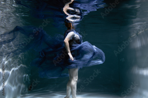 A beautiful girl in a blue dress swims underwater in the pool like a little mermaid from a fairy tale