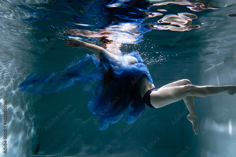 Naklejka premium A beautiful girl in a blue dress swims underwater in the pool like a little mermaid from a fairy tale
