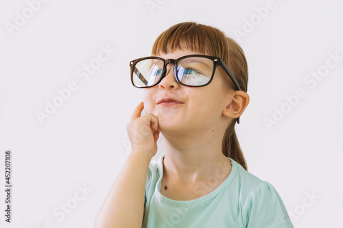 A thoughtful little girl with glasses. Cute thoughtful blonde girl standing on a white background. A beautiful child playing the thinker. The little girl is up to something. Solution of the issue