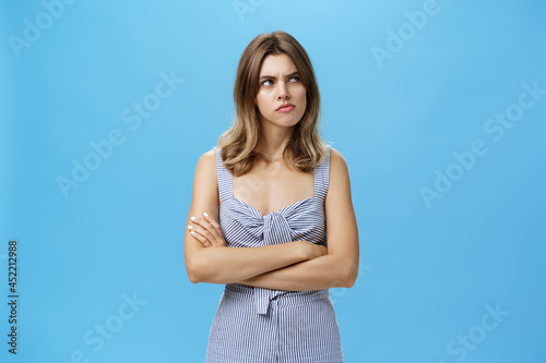 Studio shot of woman thinking seriously being suspicious and hesitant pursing lips holding hands crossed on chest looking at upper right corner with frown, thinking over blue background