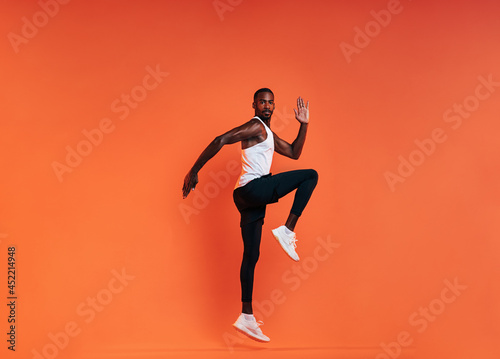 Young fit man doing exercises in studio. Male athlete jumping in the air over an orange background.