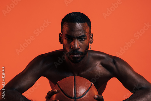 Bare-chested sportsman clutching basket ball in front of him while standing in a studio