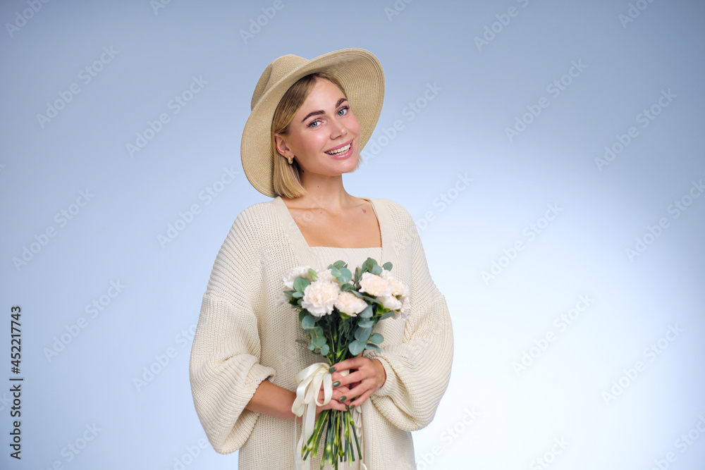 A young beautiful blonde caucasian woman with a short haircut in a beige suit and hat with bouquet of white flowers on blue background. Beauty portrait. The girl smiles, clean skin and white teeth. A young beautiful blonde caucasian woman with a short haircut in a beige suit and hat with bouquet of white flowers on blue background. Beauty portrait. The girl smiles, clean skin and white teeth.
