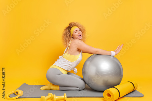 Sideways shot of curly satisfied young woman poses on knees at fitness mat keeps arms on swiss ball smiles pleasantly surrounded by headphones karemat dumbbells isolated over yellow background