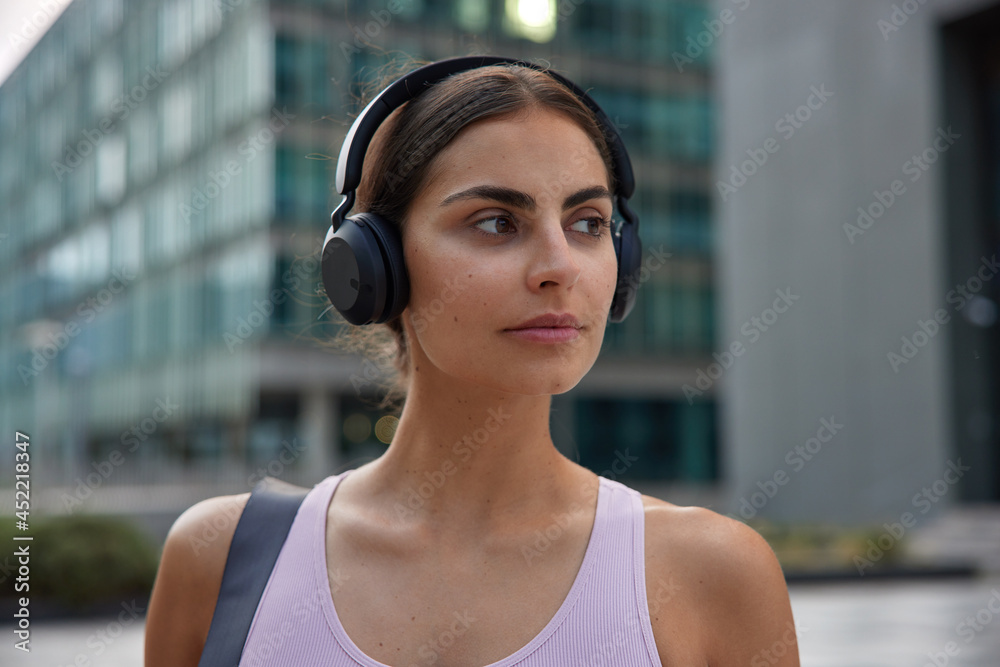 Close up shot of pensive brunette woman wears casual t shirt listens music via stereo headphones strolls in city discovers new places of interest looks away. People lifestyle and leisure concept