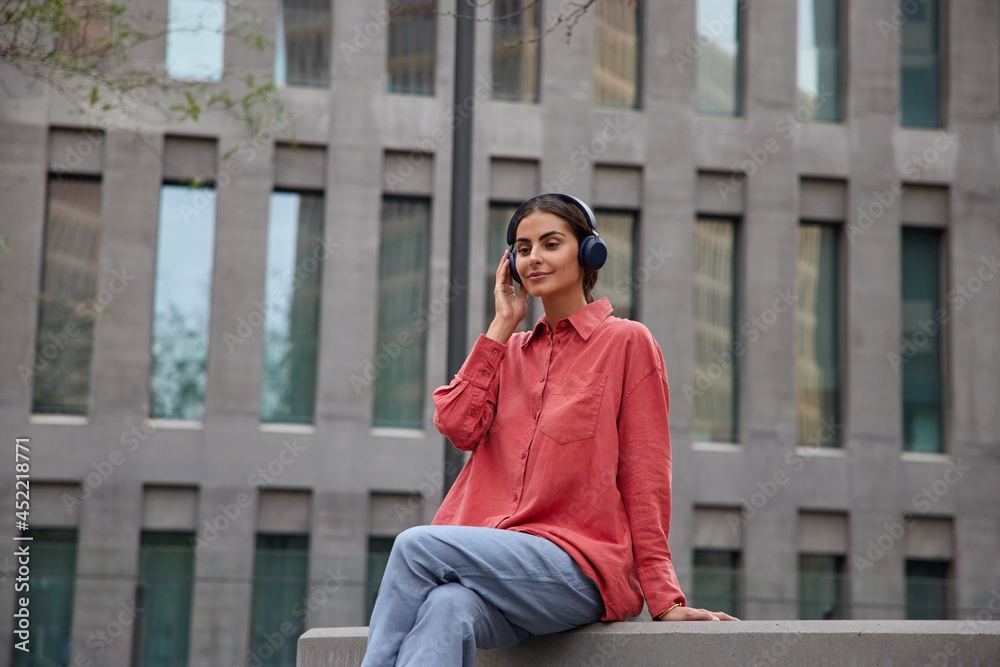 Beautiful brunette woamn sits outdoors listens music or audio podcast via wireless headphones enjoys leisure pastime dressed in red shirt and trousers takes break after strolling. Lifestyle concept