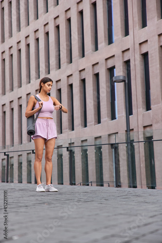 Young woman does sport outdoor checks time on smartwatch carries rolled yoga mat dressed in sportswear waits for someone poses near modern building gets ready to start. Athletic sportswoman.