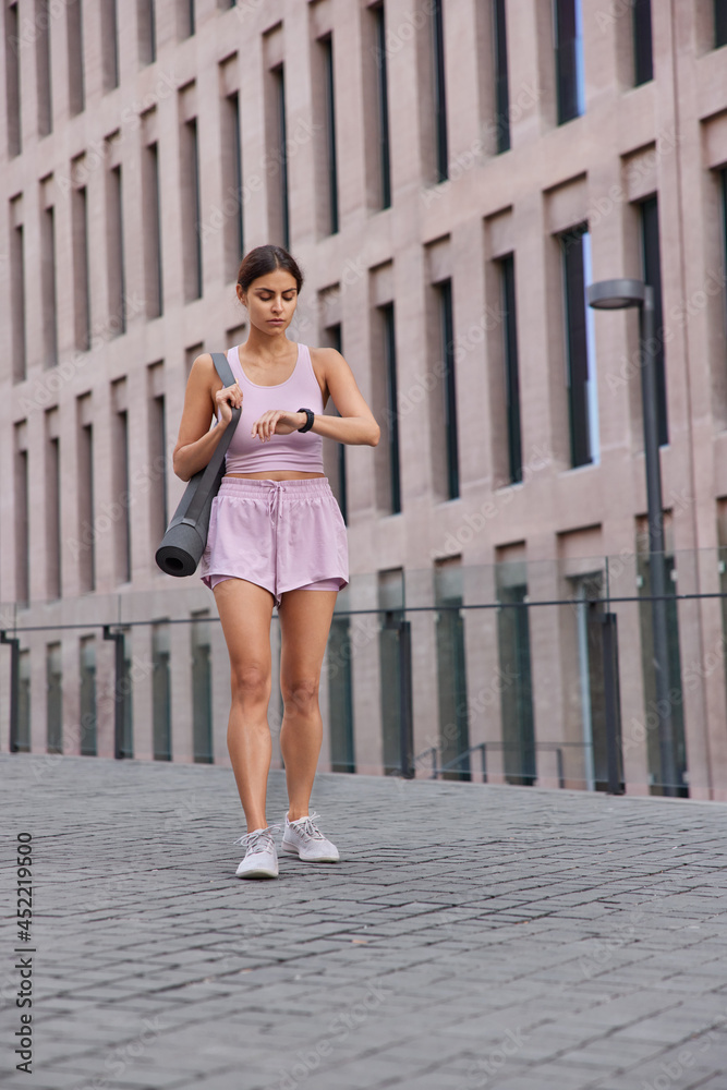 Vertical shot of Caucasian sportswoman walks outdoor in sportswear looks at modern wearable smartwatch tracking time checks how much calories she burnt after cardio training poses in urban place Vertical shot of Caucasian sportswoman walks outdoor in sportswear looks at modern wearable smartwatch tracking time checks how much calories she burnt after cardio training poses in urban place