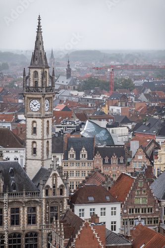 Gent, Flanders, Belgium - July 30, 2021: Slender postal clock tower over dense cityscape in the rain with tall red crane and green hills in back. Abundance of medieval facades and gables.