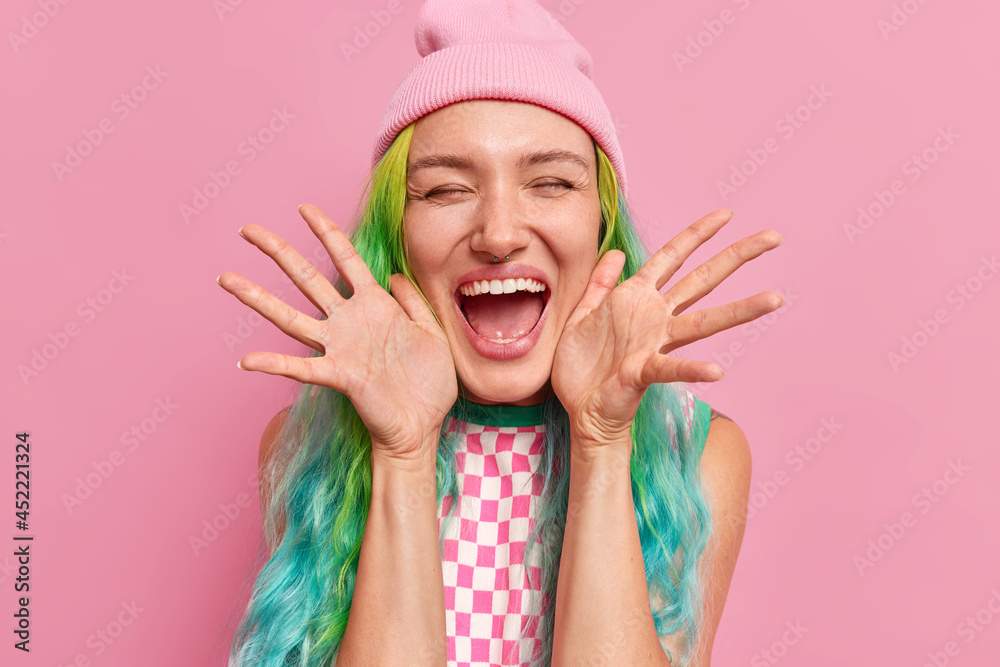 Photo of overjoyed woman keeps palms sideways near face laughs out happily feels very happy keeps eyes closed widely opened mouth wears hat and dress isolated over pink background. Emotions concept Photo of overjoyed woman keeps palms sideways near face laughs out happily feels very happy keeps eyes closed widely opened mouth wears hat and dress isolated over pink background. Emotions concept