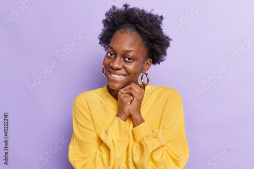 Portrait of good looking dark skinned young woman with natural curly hair keeps hands together near face smiles toothily feels touched wears casual yellow jumper isolated over purple background.