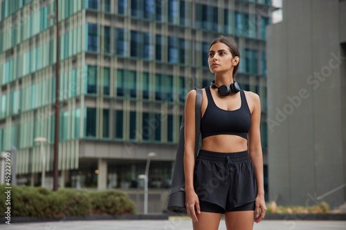 Sport and recreation concept. Self confident slim sportswoman in black cropped top and shorts carries karemat strolls outdoor in downtown leads healthy lifestyle has regular training in open air