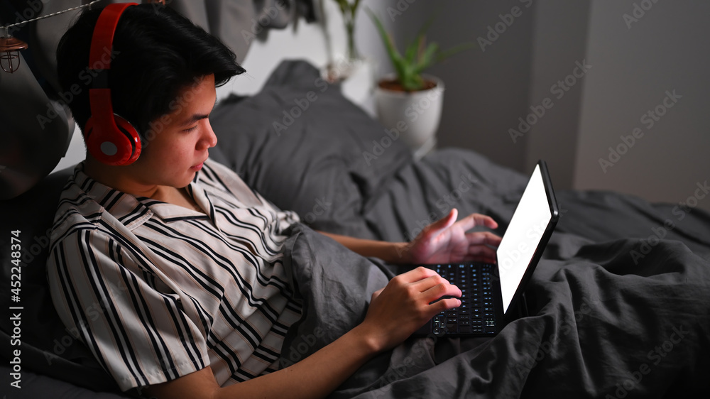 Young Asian man wearing headphone and browsing internet with computer tablet on his bed.
