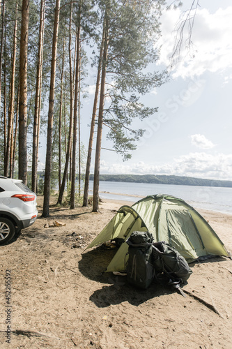 A green tent on the river bank. Sandy beach and pine forest on a summer day. there is a car in the background. Camping and outdoor recreation.