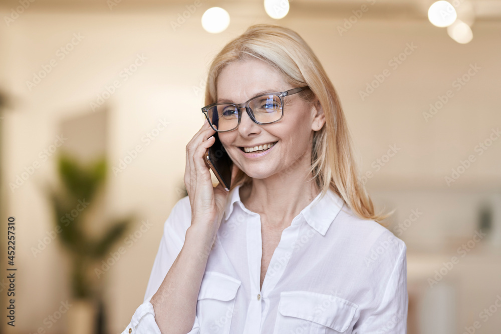Pleasant talk. Attractive mature woman with blonde hair and glasses in casual wear talking on her phone, standing at home Pleasant talk. Attractive mature woman with blonde hair and glasses in casual wear talking on her phone, standing at home