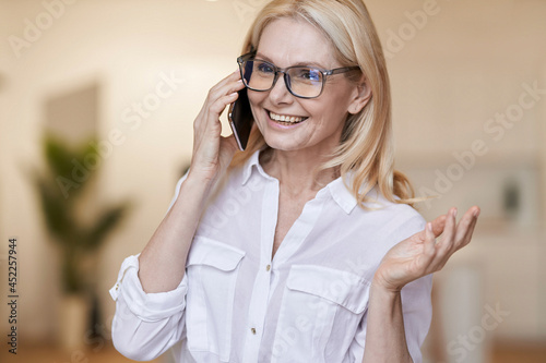 Smiling mature woman with blonde hair and glasses in white shirt talking on her phone, standing at home