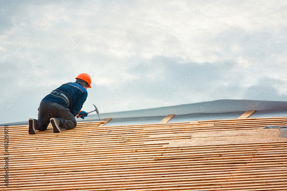 Worker hammer nails in wooden board on roof at height. Roofer carpenter works on roof in hardhat and safety harness. Renovation, repair and fixing frame work house. New building construction Worker hammer nails in wooden board on roof at height. Roofer carpenter works on roof in hardhat and safety harness. Renovation, repair and fixing frame work house. New building construction