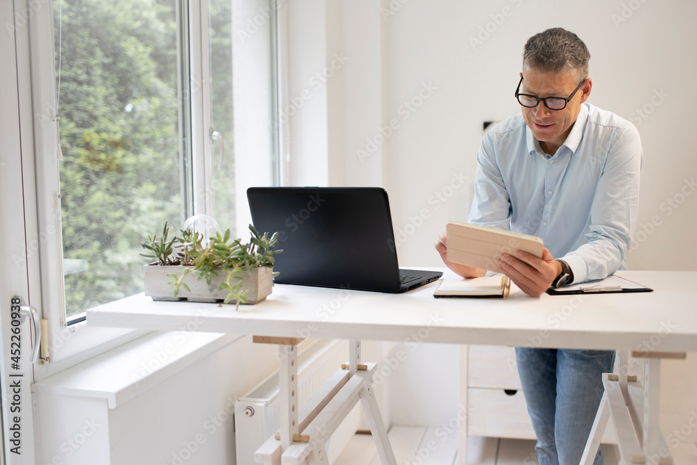 business man with blue shirt and black glasses is standing behind standing table and is working with his tablet and a black laptop in a modern office